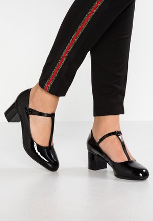 Voeten in zwarte lak T-strap blokhak schoenen gecombineerd met zwarte broek met rode en gouden zijstrepen.