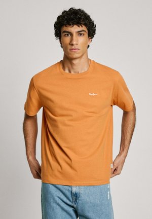 Camiseta de algodón naranja con cuello redondo, mangas cortas y un pequeño logo blanco en el pecho. Llevada con jeans de color azul claro.