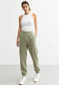 Witte mouwloze top, olijfgroene sweatpants met elastische tailleband en boorden, gecombineerd met witte sneakers, staande op een lichte vloer.