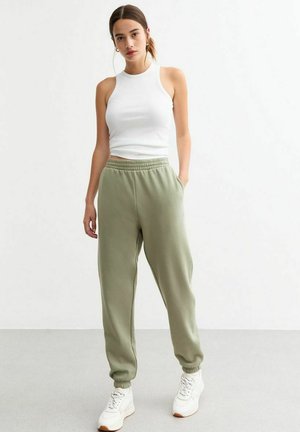 Witte mouwloze top, olijfgroene sweatpants met elastische tailleband en boorden, gecombineerd met witte sneakers, staande op een lichte vloer.