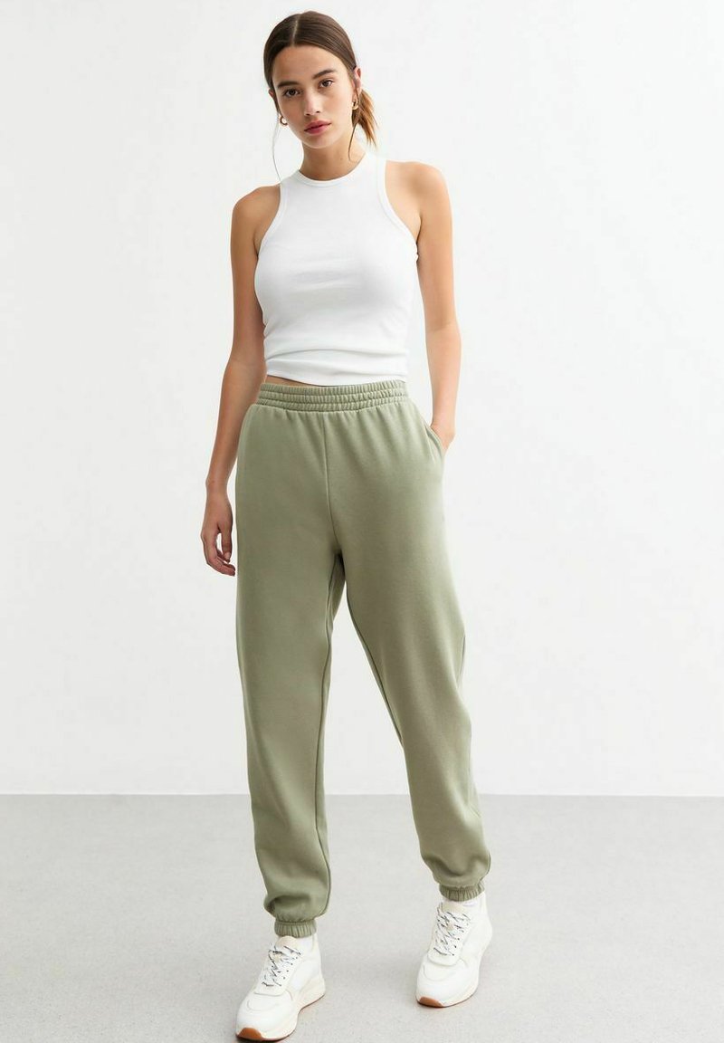 Witte mouwloze top, olijfgroene sweatpants met elastische tailleband en boorden, gecombineerd met witte sneakers, staande op een lichte vloer.