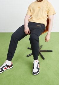 Person som bär en beige Nike-t-shirt, svarta cargo-joggers och svarta, vita och röda Nike-sneakers, sitter på en modern orange stol på en grön matta.