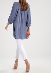 Tunica azzurra chiara a maniche lunghe con schiena plissettata, abbinata a jeans bianchi arrotolati e tacchi alti color nude, che mostrano una silhouette rilassata.