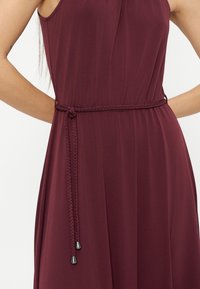 Φόρεμα μπορντό χωρίς μανίκια με πλεκτή ζώνη στη μέση, δεμένη στο πλάι, που αποκαλύπτει το μέσο του σώματος και μέρος των χεριών, σε απλό φόντο.