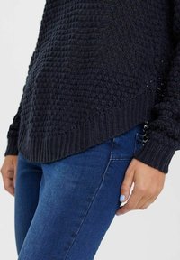 Maglione lavorato a maglia di un blu navy scuro con corpo testurizzato e polsini a coste, abbinato a jeans blu aderenti. Il maglione presenta un orlo con una leggera incurvatura.