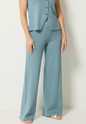 Mujer con pantalones anchos de color azul claro y top sin mangas con botones a juego, con sandalias de punta abierta, de pie frente a un fondo liso.