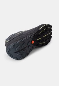 Black running shoe sole with aggressive traction pattern. Features textured rubber, a midsole with yellow accents, and a distinctive red logo.