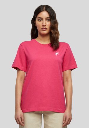 Femme aux cheveux mi-longs ondulés et foncés, portant un T-shirt rose vif avec un petit cœur blanc et des rayons sur la poitrine gauche.