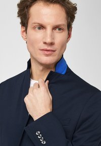 Mann, der den Kragen eines marineblauen Blazers mit blauer Innenseite anpasst, trägt ein weißes Hemd darunter und schaut leicht zur Seite vor einem schlichten Hintergrund.