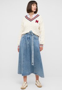 Kremowy sweter w warkoczowy wzór z czerwonym i granatowym dekoltem w serek, z literą "M". Spódnica z wysokim stanem z niebieskiego denimu z paskiem w talii oraz białe sneakersy.