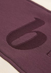 Bordo kangas, millel on pehme tekstuur, kannab tumedamas toonnis broderdatud suurt numbrit "0", mis on keskendatud materjalile.