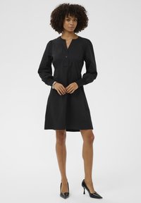 Fekete ruha gombos kivágással, hosszú puffos ujjakkal és bővülő szoknyával. Illeszkedő mandzsettagombokkal és sima felülettel rendelkezik.