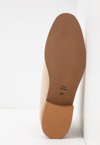 Semelle de chaussure en daim beige avec talon en caoutchouc marron, taille 37, tamponnée "Made in Portugal", reposant contre un mur blanc d'angle.