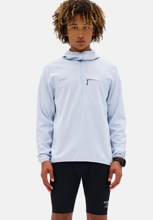 Giovane con capelli ricci che indossa un pullover azzurro chiaro con zip a quarto e pantaloncini sportivi neri, in piedi davanti a uno sfondo bianco semplice.