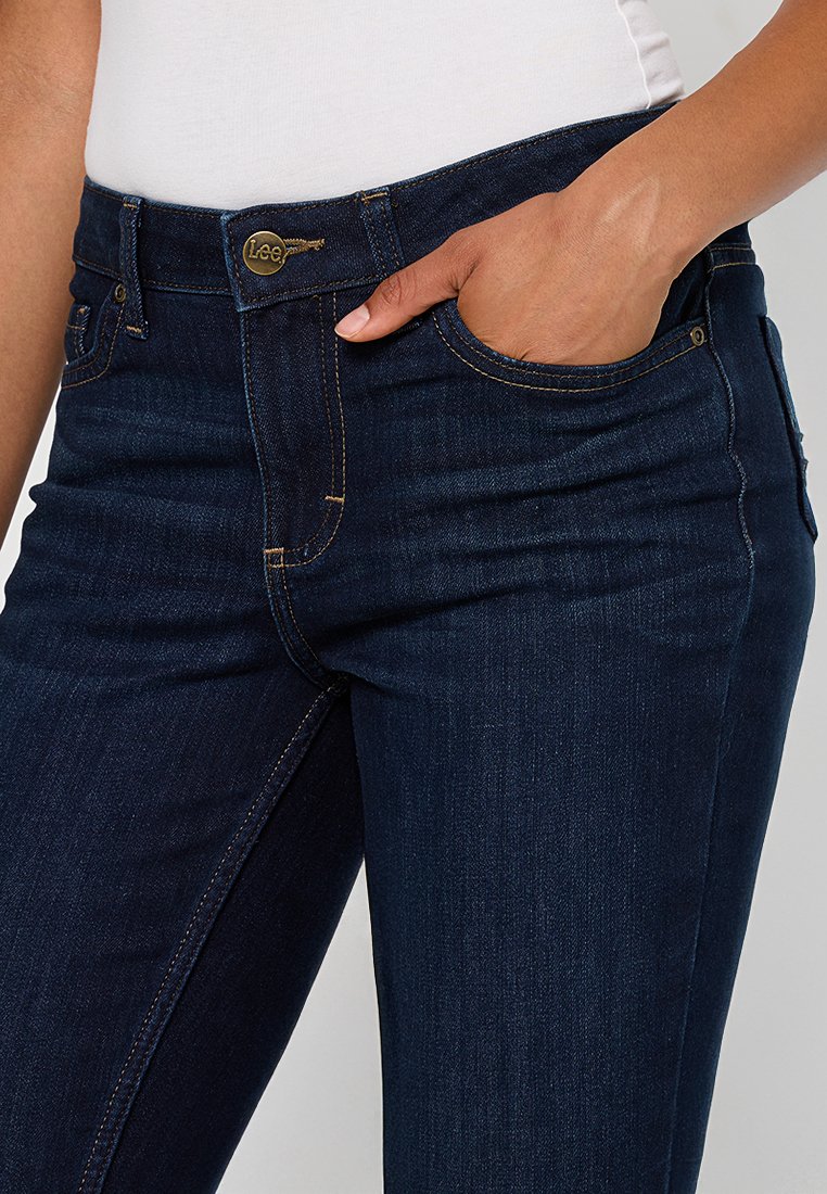 Jeans in denim blu scuro con vita a media altezza, dettagli in metallo dorato e tasche frontali. Le cuciture visibili evidenziano la vestibilità sartoriale.