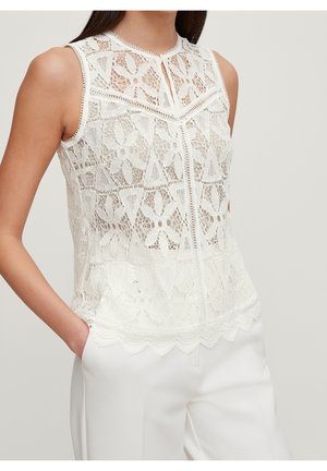 Sleeveless white lace top with floral and geometric patterns, scalloped hem, and keyhole neckline, worn with white tailored pants.