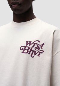Cremefarvet sweatshirt med et iøjnefaldende broderet bordeaux logo, der læser "Wrst Bhvr" på venstre bryst. Klassisk crewneck-design.