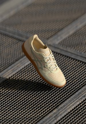 Beżowo - jasnobrązowy sneaker ze sznurowaniem, z markowymi paskami po bokach, spoczywający na teksturowanej metalowej kratce.