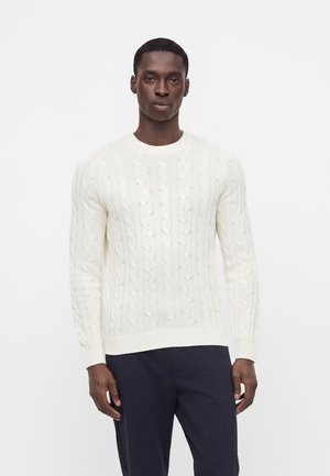 Homme portant un pull en maille torsadée blanc et un pantalon foncé, debout sur un fond blanc uni.