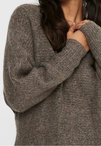 Mujer vestida con un suéter de punto marrón texturizado y mangas abullonadas, sosteniendo el cuello cerca de su cuello con ambas manos.