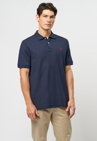 Bărbat purtând un tricou polo albastru închis cu un mic logo roșu pe piept și pantaloni bej, stând în fața unui fundal simplu, deschis la culoare.