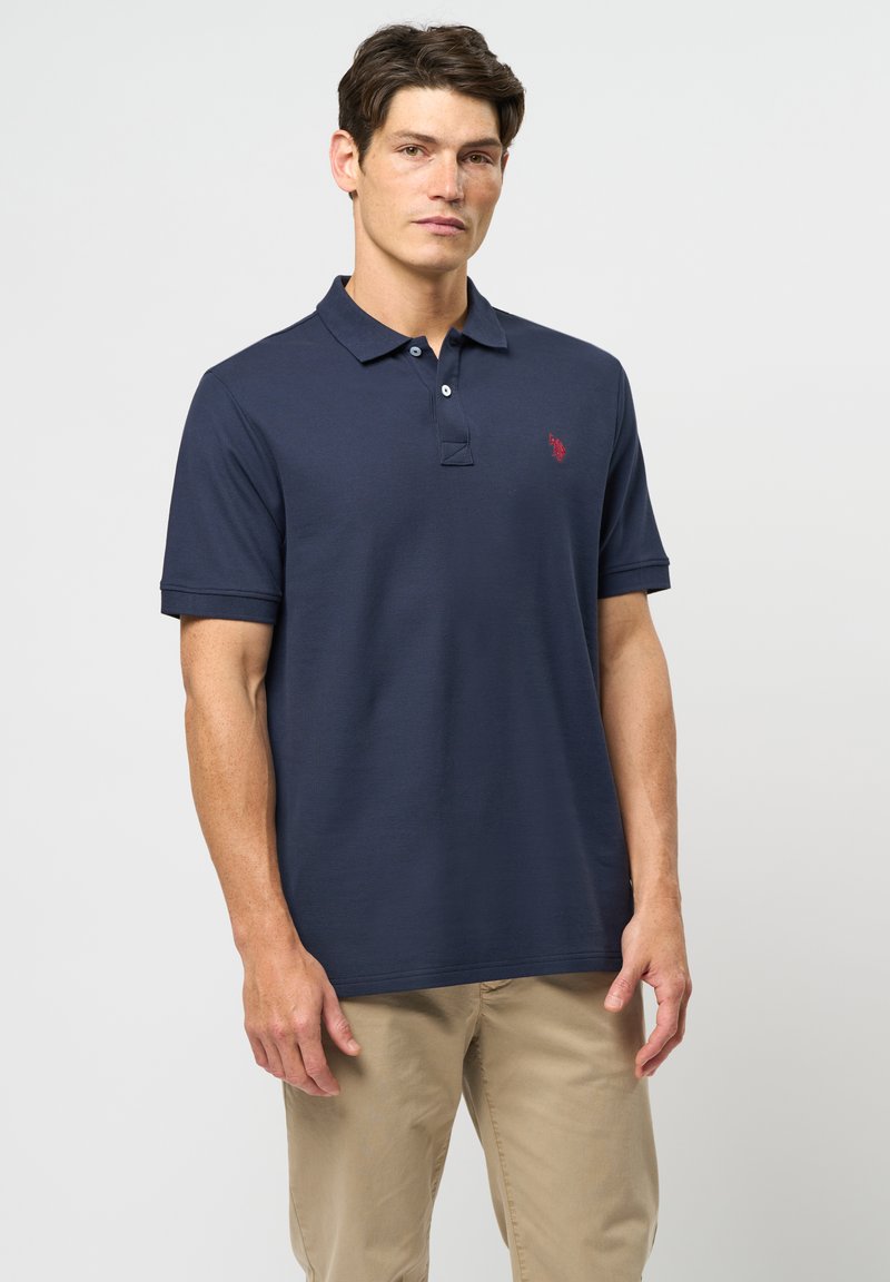 Bărbat purtând un tricou polo albastru închis cu un mic logo roșu pe piept și pantaloni bej, stând în fața unui fundal simplu, deschis la culoare.