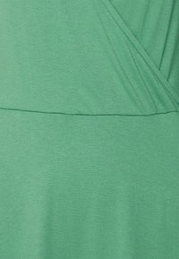 Tecido verde, leve e texturizado, com um design cruzado. Costura horizontal a meio. Aparência suave e casual.