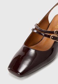 Brown patent leather Mary Jane shoe with a square toe, two slim straps with gold-tone buckles, and a smooth finish.