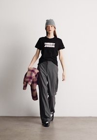 Μαύρη μπλούζα τύπου T-shirt με λευκό λογότυπο "TOMMY JEANS", γκρι σκουφάκι, γκρι παντελόνι με ρίγες, κρατώντας ένα μωβ καρό σακάκι, μαύρα παπούτσια.