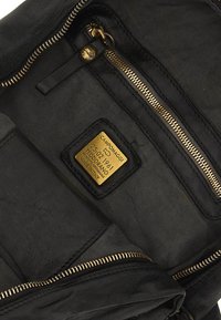 Interno di una borsa in pelle nera con una zip dorata, foderato in tessuto e un'etichetta dorata con nome del marchio e dettagli. Due tasche visibili.