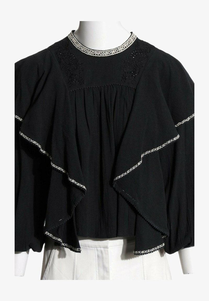 Blouse noire à manches longues avec des couches à volants et des bords brodés blancs sur le col, les manches et les volants, portée avec un pantalon blanc.