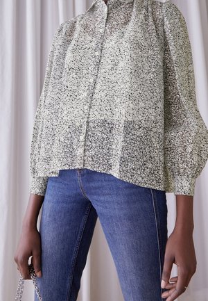 Osoba ubrana w jasnoszarą, kwiatową bluzkę na guziki z bufkami na rękawach i niebieskie dżinsy, trzymająca srebrną torebkę na łańcuszku na tle białej zasłony.