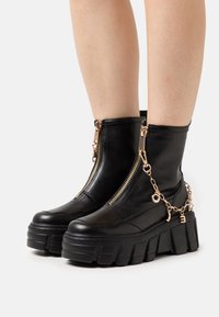 Mustat nilkkureita, jotka on valmistettu sileästä nahasta. Varustettu paksulla pohjalla, kultaisilla vetoketjuyksityiskohdilla ja koristeellisia ketjuja riipuksilla.