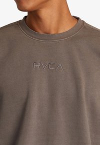 Sweat-shirt marron avec un col rond et des détails côtelés, arborant un logo brodé "RVCA" en fil assorti sur la poitrine.