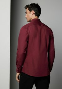 Cămașă lungă călcată, de culoare vișinie, cu un design ajustat, manșete cu nasturi și o textură netedă. Vederea din spate arată un tiv curbat și o croială subtilă.