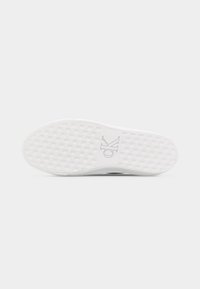 Semelle d'une basket blanche avec un motif tissé et le logo "ck" embossé au centre sur un fond gris clair uni.