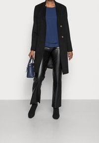 Long manteau noir par-dessus un haut bleu marine, assorti à un pantalon en fausse cuir noir brillant et des bottines noires. Un petit sac à main bleu marine est tenu à la main.