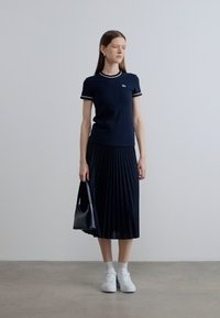 Μπλε ναυτικό πόλο μπλουζάκι με λευκή ριγέ γιακά, σε συνδυασμό με πλισέ μίντι φούστα. Το μοντέλο κρατάει μια μαύρη τσάντα και φοράει λευκά αθλητικά παπούτσια.