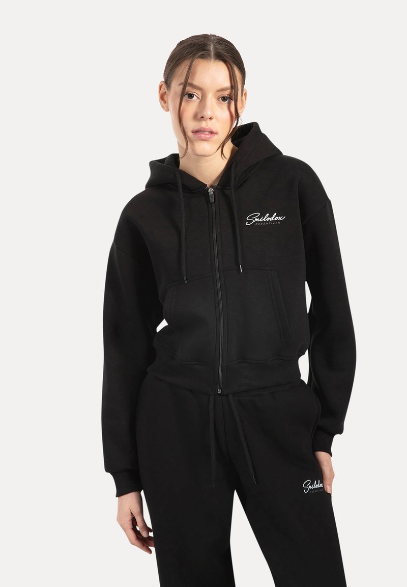 Hoodie noir zippé en matériau doux, avec une poche avant et des cordons de serrage. Associé à un pantalon de survêtement noir assorti.