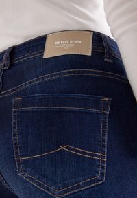 Granatowe dżinsy z denimu z brązową skórzaną łatą z napisem "KOCHAMY DENIM" nad tylną kieszenią. Szycie w kontrastującym kolorze.