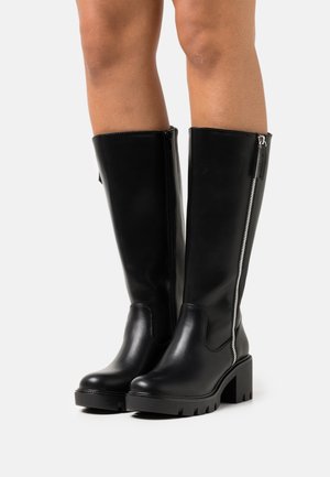 Černé kožené kozačky ke kolenům s masivními podpatky a stříbrnými bočními zipy, nošené na holých nohou na jednobarevném pozadí.