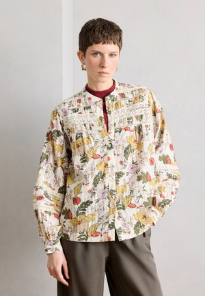 Osoba s krátkymi hnedými vlasmi, ktorá má na sebe kvetinovú blúzku s dlhým rukávom a čipkovými detailmi a olivové nohavice, stojaca pri jednoduchom svetlom múre.