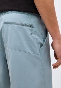 Κοντινό πλάνο ατόμου που φοράει ανοιχτό μπλε σορτς με φερμουάρ στην πίσω τσέπη και πλευρική ραφή, μαύρο πουκάμισο, και το χέρι χαλαρά στο πλάι.