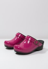 Pink patent clogs with a rounded toe, a black strap and buckle, and a slight heel. The surface is glossy and smooth with visible stitching.
