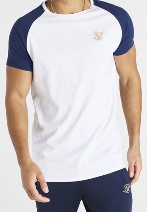 Man draagt een wit t-shirt met donkerblauwe mouwen en donkerblauwe broek, beide met een klein goudlogo aan de linkerzijde.