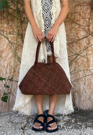 Persoon in een jurk met patroon en een omslagdoek met franjes houdt een grote bruine gevlochten handtas vast, draagt zwarte sandalen en staat op grind met een muur bedekt met wijnstokken op de achtergrond.