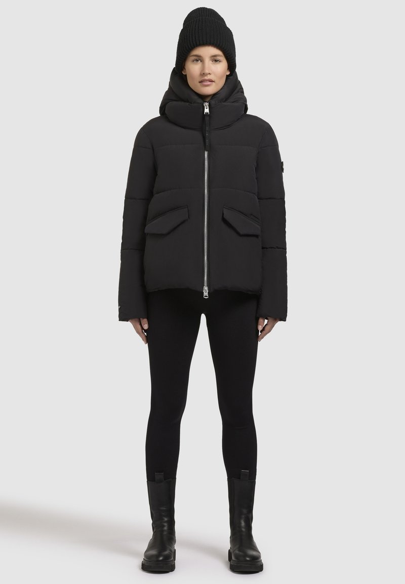 Frau in einer schwarzen Steppjacke mit vorderen Taschen, schwarzen Leggings, schwarzen Stiefeln und einer schwarzen Strickmütze, die vor einem weißen Hintergrund steht.
