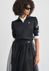 Μαύρο μπουφάν με φερμουάρ, με λευκές ρίγες και λογότυπο, σε συνδυασμό με φούστα από μαύρο τούλι. Το μπουφάν έχει φουσκωτά μανίκια και εφαρμοστή μέση.