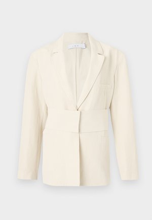 Personne à la peau foncée portant un blazer crème oversize, un pantalon crème à jambes larges, une chemise blanche col V et des chaussures noires, debout sur fond blanc.