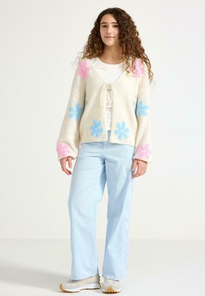 Cardigan crème moelleux à motifs floraux roses et bleus, fermeture à nouer devant, associé à un jean bleu clair et des baskets blanches.